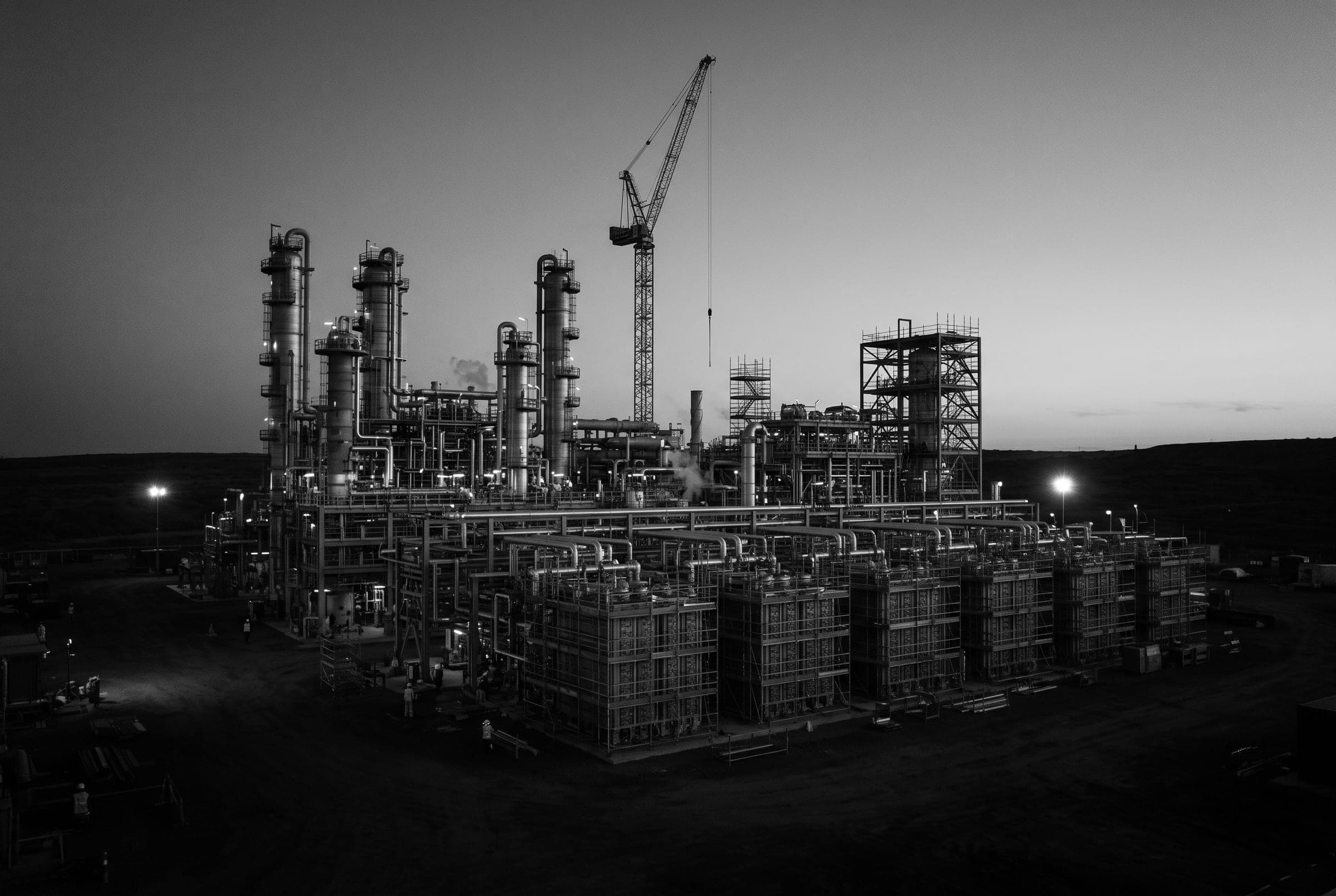 Unité industrielle en construction au crépuscule : colonnes de procédé, échafaudages et grue.