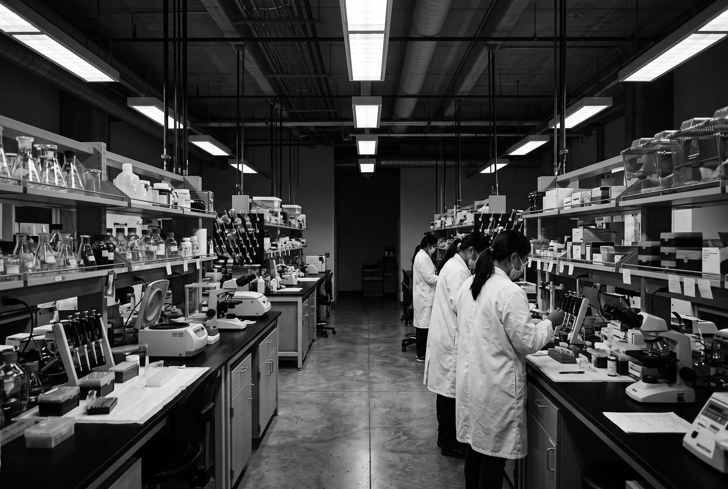 Laboratoire de recherche : chercheurs en blouse blanche aux paillasses, sous des rampes lumineuses.